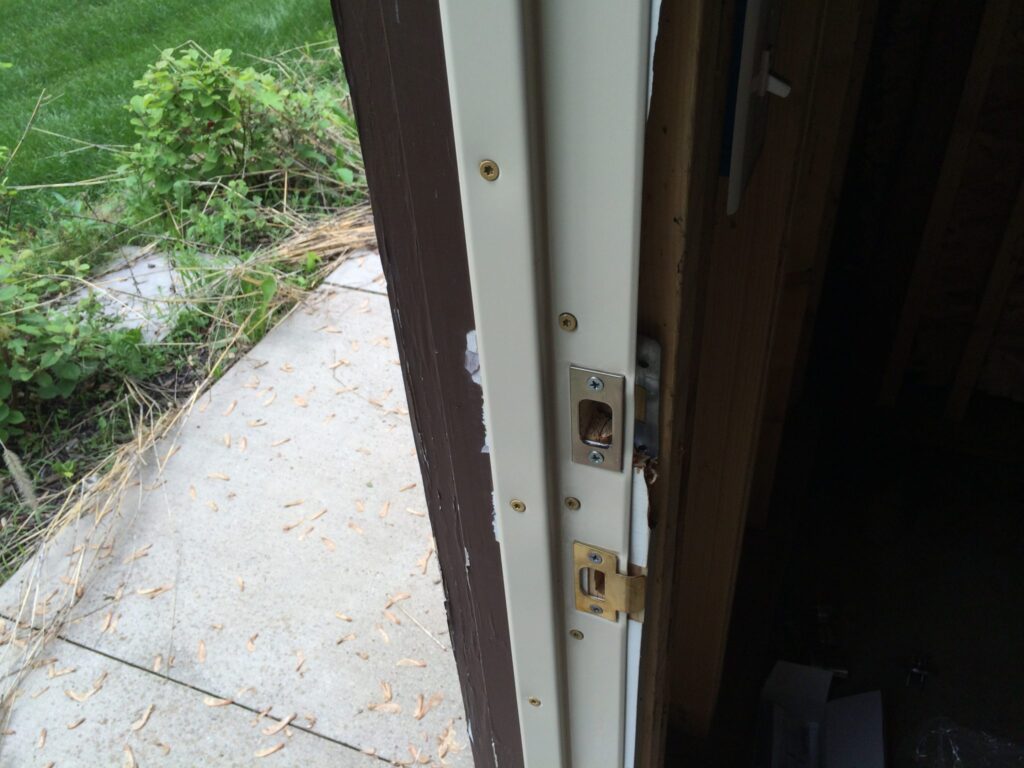 Close-up of a damaged door frame with a broken lock, shown from an indoor perspective with foliage and a pathway outside.